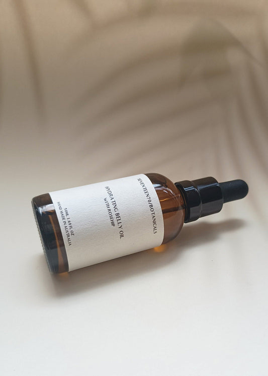 A bottle of Hydrating Belly Oil with Rosehip, displaying a clear amber liquid in a dark glass bottle with a black dropper. The label is white with text.