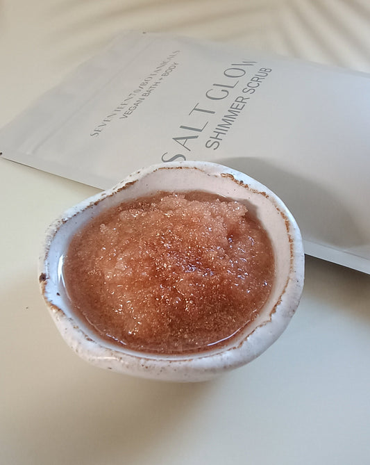 A pouch of Salt Glow Shimmer Scrub with a small container filled with the pinkish scrub powder beside it.