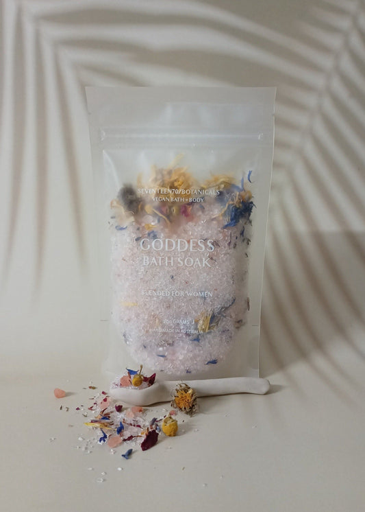 A sealed pouch labeled 'Goddess Bath Soak' with a shower of flower petals and bath salts spilled in front of it, set against a white background with a shadow cast over it.