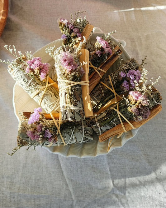 Bundles of sage and palo santo stick with flowers on a white plate.