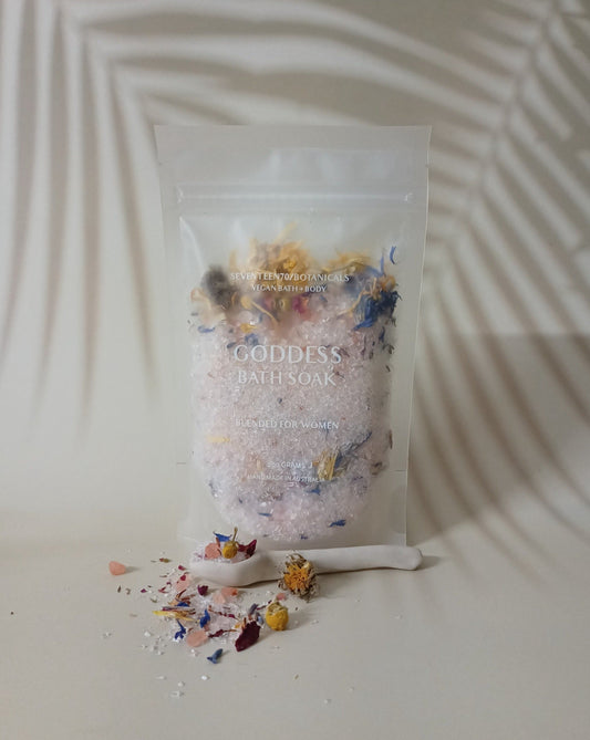 A sealed pouch labeled 'Goddess Bath Soak' with a shower of flower petals and bath salts spilled in front of it, set against a white background with a shadow cast over it.